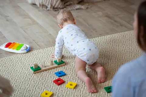 Baby crawling towards a wooden shape sorter while a carer watches