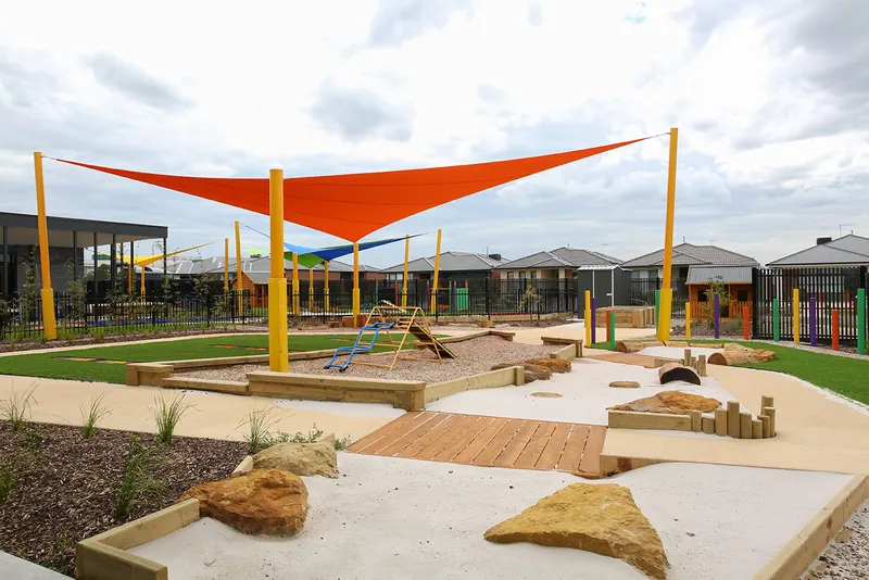 Natural playground with sandpit, timber decking and boulders under an orange shade sail