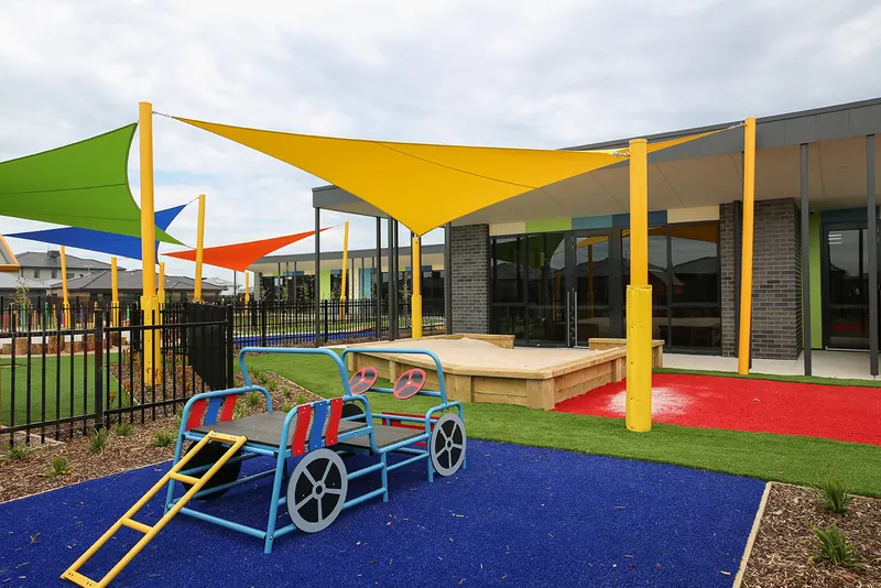 Outdoor play area with colourful shade sails, soft-fall surfaces and a climbing car