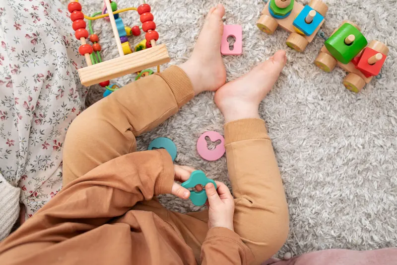 Toddler sitting on a rug playing with wooden shape and train toys